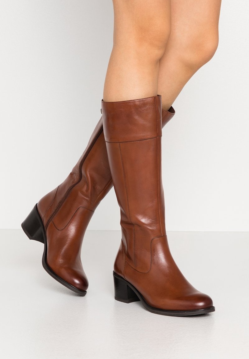 Bottes en cuir marron montant jusqu'au genou avec talons épais de hauteur moyenne, portées sur des jambes nues sur un fond clair uni.