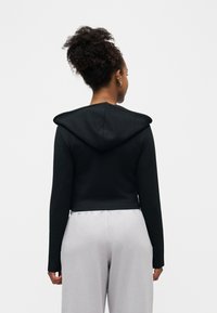 Femme aux cheveux bouclés attachés en chignon, portant un sweat à capuche noir court et un pantalon gris clair, debout de dos sur un fond blanc uni.