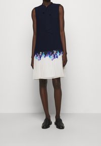 Robe sans manches avec un corsage bleu marine et une jupe plissée crème ornée de motifs floraux violets à l'ourlet. Chaussures noires sans lacets.