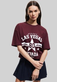 T-paita, jossa on marooni puuvilla ja valkoinen grafiikka, jossa lukee "LAS VEGAS 80 NEVADA" tähtikuvion kera. Yhdistetty siniseen, laskostettuun mini-hameeseen.