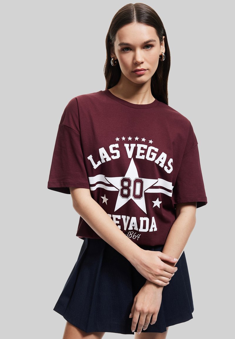 T-paita, jossa on marooni puuvilla ja valkoinen grafiikka, jossa lukee "LAS VEGAS 80 NEVADA" tähtikuvion kera. Yhdistetty siniseen, laskostettuun mini-hameeseen.