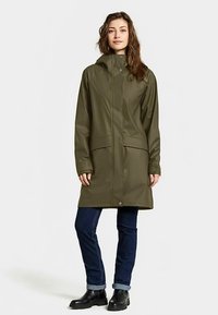 Waterdichte olijfgroene jas met capuchon, drukknoopsluiting aan de voorkant, twee grote zakken, lange mouwen en een lengte tot halverwege de dij. Draagt bij donkere spijkerbroek en laarzen.