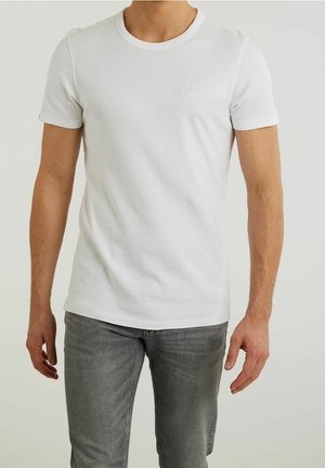 Mężczyzna w prostym białym t-shircie z krótkim rękawem i szarych dżinsach stojący na jasnym, jednolitym tle.