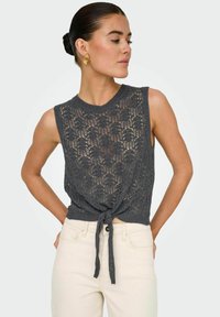 Haut court en tricot gris avec un motif en dentelle, sans manches, et un détail à nouer à la taille, associé à un pantalon clair.