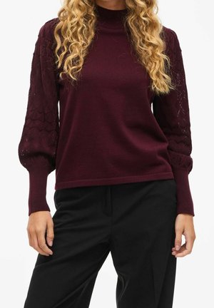 Femme portant un pull bordeaux avec des manches ballon texturées et un pantalon noir, debout devant un fond blanc.