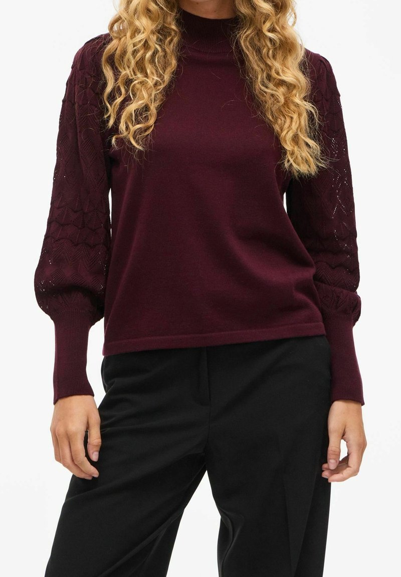 Femme portant un pull bordeaux avec des manches ballon texturées et un pantalon noir, debout devant un fond blanc.