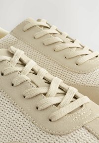 Zapatillas beige que combinan malla texturizada y cuero suave, con cordones planos tejidos y un diseño de punta redondeada.