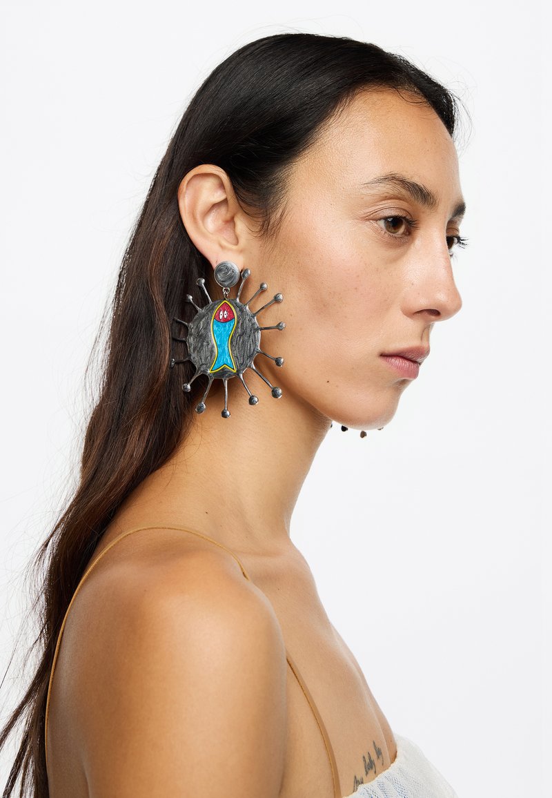 Pendientes grandes y coloridos con un diseño de pez azul sobre un fondo metálico texturizado, con acentos circulares y detalles colgantes.