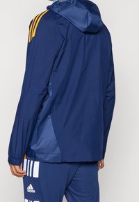 Marine windbreaker jack met een capuchon, gestructureerde mouwen en oranje drie-streepdesign details. Elastische manchetten; lichtgewicht stof.