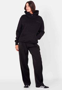 Zwarte capuchontrui met een kangoeroezak, gecombineerd met loszittende zwarte broeken. De stof oogt zacht, met een gladde textuur en een minimalistisch ontwerp.