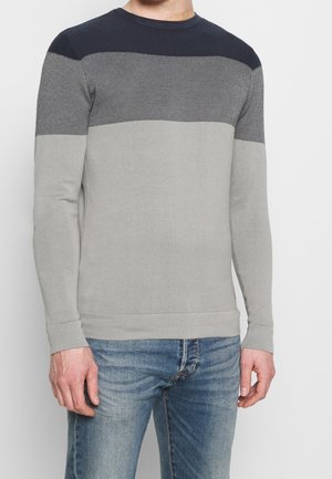 Homme portant un pull à manches longues et col rond avec des blocs de couleur horizontaux bleu foncé, gris et gris clair, associé à un jean bleu.