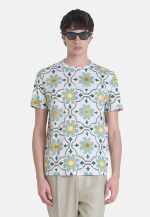 Camisa de manga corta hecha de tejido ligero con un patrón geométrico floral en amarillo, azul verdoso y gris sobre un fondo blanco.