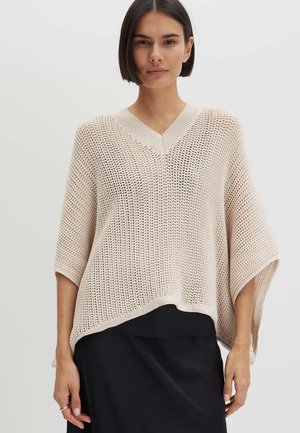 Vrouw met steil donker haar, die een losse, lichtbeige gebreide trui met V-hals en een zwarte rok draagt tegen een effen achtergrond.