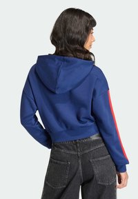 Gekürzter blauer Hoodie mit Kapuze, versehen mit roten Seitenstreifen. Das Material wirkt weich und glatt. Kombiniert mit dunklen Jeanshosen.