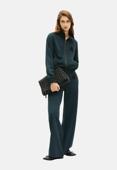 Mørkegrønt tracksuit med jakke med lynlås og matchende bukser, der har et struktureret blomsteremblem. Sort quiltet clutch-taske med lynlås.