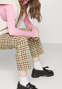 Eng anliegendes rosa Langarmshirt, cremefarbenes Oberteil, grün-braune karierte kurze Hosen, schwarze Schuhe mit Quastenakzent und weiße Socken.