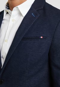 Marinblå blazer i texturerad tyg, med enkelknappsstängning och smal passform. Har en bröstficka med detaljer i tre färger.