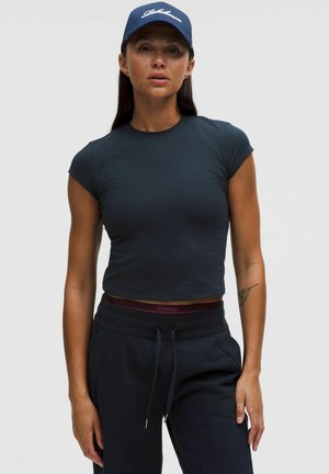 Femeie purtând tricou scurt bleumarin, pantaloni sport bleumarin și șapcă bleumarin cu inscripția "Lululemon", pe un fundal simplu, deschis.