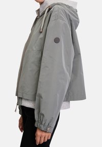 Lichtgrijze cropped jas met capuchon, elastische manchetten en voorzakken. De gestructureerde stof heeft een grijze binnenvoering en een logo-accent.