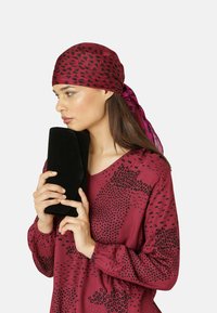 Rote Bluse mit geometrischem schwarzen Muster, kombiniert mit einem kastanienbraunen Schal und einer schwarzen Clutch, die mit beiden Händen gehalten wird. Sanfte Texturen sind zu sehen.