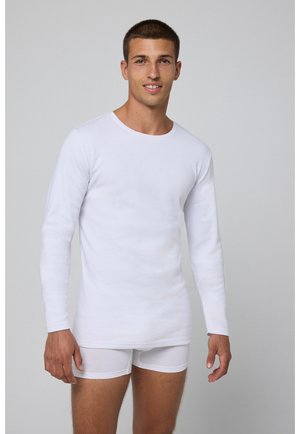 Camiseta térmica larga blanca de manga larga, hecha de algodón, con cuello redondo. Combinada con calzoncillos boxers blancos, creando un look de ropa interior en capas.