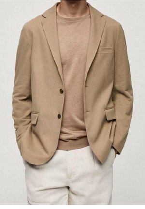 Beige blazer with two front pockets and brown buttons, paired with a light brown sweater. White trousers complete the outfit. Smooth texture throughout.