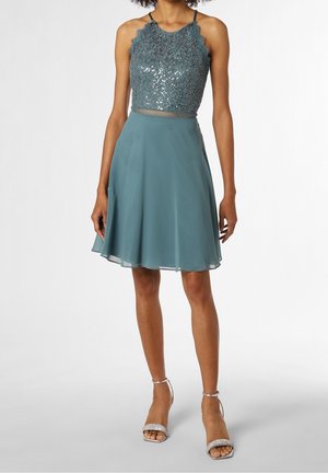 Frau trägt ein ärmelloses türkisfarbenes Kleid mit Spitze und Pailletten am Oberteil sowie einem fließenden Chiffonrock, kombiniert mit silbernen High-Heel-Sandalen.