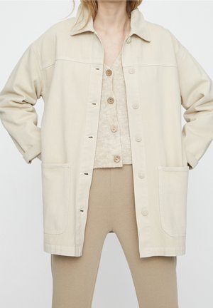 Person wearing beige buttoned cardigan, loose off-white jacket with pockets, and beige pants, standing with hands on hips.