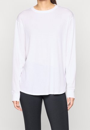 Woman wearing plain white long-sleeve shirt and black leggings, standing against a light neutral background.