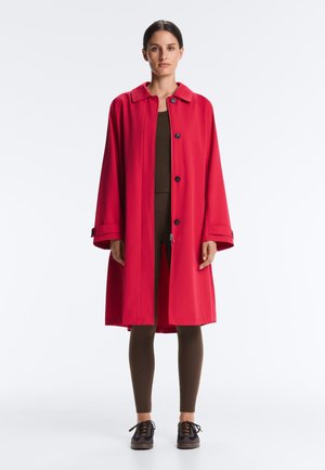 Nő áll egy élénkpiros, térdig érő kabátban, sötétbarna leggings és ing fölött, fekete sportcipővel, fehér háttér előtt.