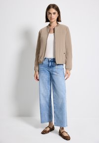 Femme se tenant contre un fond blanc, portant une veste beige zippée, un haut blanc, un jean large bleu clair et des ballerines à imprimé léopard.