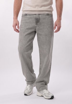 Mann trägt hellgraue locker sitzende Jeans und weiße Sportschuhe, steht vor einem einfarbigen Hintergrund.