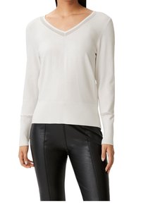 Lekki, biały sweter z dekoltem w serek i długimi rękawami. Posiada ściągacze na mankietach i dole oraz przezroczyste detale przy dekolcie. Połączony z czarnymi spodniami o wyglądzie skóry.