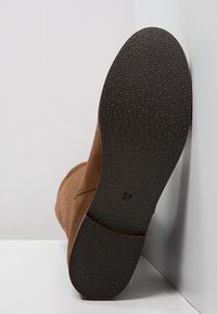 Bottine en daim marron avec une semelle en caoutchouc noire texturée, inclinée sur un fond blanc. Taille indiquée comme 37 sur la semelle.