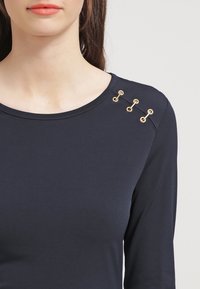 Marinefarbener Langarmshirt mit Rundhalsausschnitt, das drei goldene Dekoschleifen an der Schulter aufweist. Glatte, taillierte Stoffqualität.