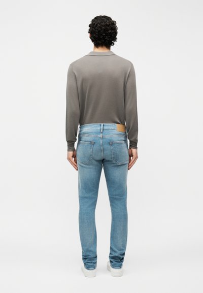 Άνδρας με σγουρά μαλλιά, ντυμένος με γκρι μακρυμάνικο πουκάμισο, ανοιχτόχρωμα μπλε τζιν και λευκά sneakers, στέκεται ανάποδα σε ένα απλό φόντο.