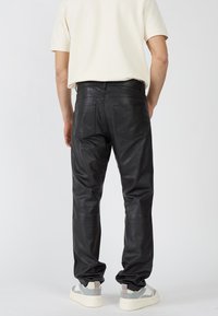 Schwarze Lederhosen mit schmalem Schnitt, ausgestattet mit zwei Gesäßtaschen und einer glatten Textur. Getragen mit einem hellen Hemd und Sneakers.