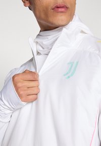 Lekka biała kurtka z mikrozamkiem, z miętowozielonym logo i akcentami kolorystycznymi na ramieniu; gładka tekstura, dopasowany krój.
