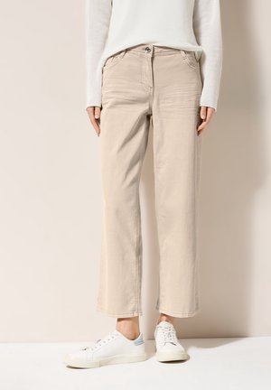 Vrouw die beige rechte broek, witte longsleeve trui en witte sneakers draagt, staand tegen een effen lichte achtergrond.