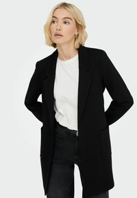 Svart strukturblazer med en uppbyggd design, notch-kragar, långa ärmar och två fickor, tillsammans med en vit t-shirt och mörka jeans.