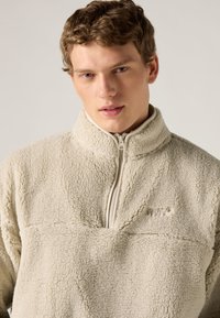 Πουλόβερ από φλις σε κρεμ απόχρωση με γιακά μισό φερμουάρ, διαθέτοντας μαλακή, υφή και ραμμένες λεπτομέρειες στο μπροστινό μέρος.