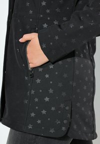 Ruka v zipové kapse černé bundy s tmavým hvězdným vzorem a patentkami blízko lemu, nošená s tmavými kalhotami.