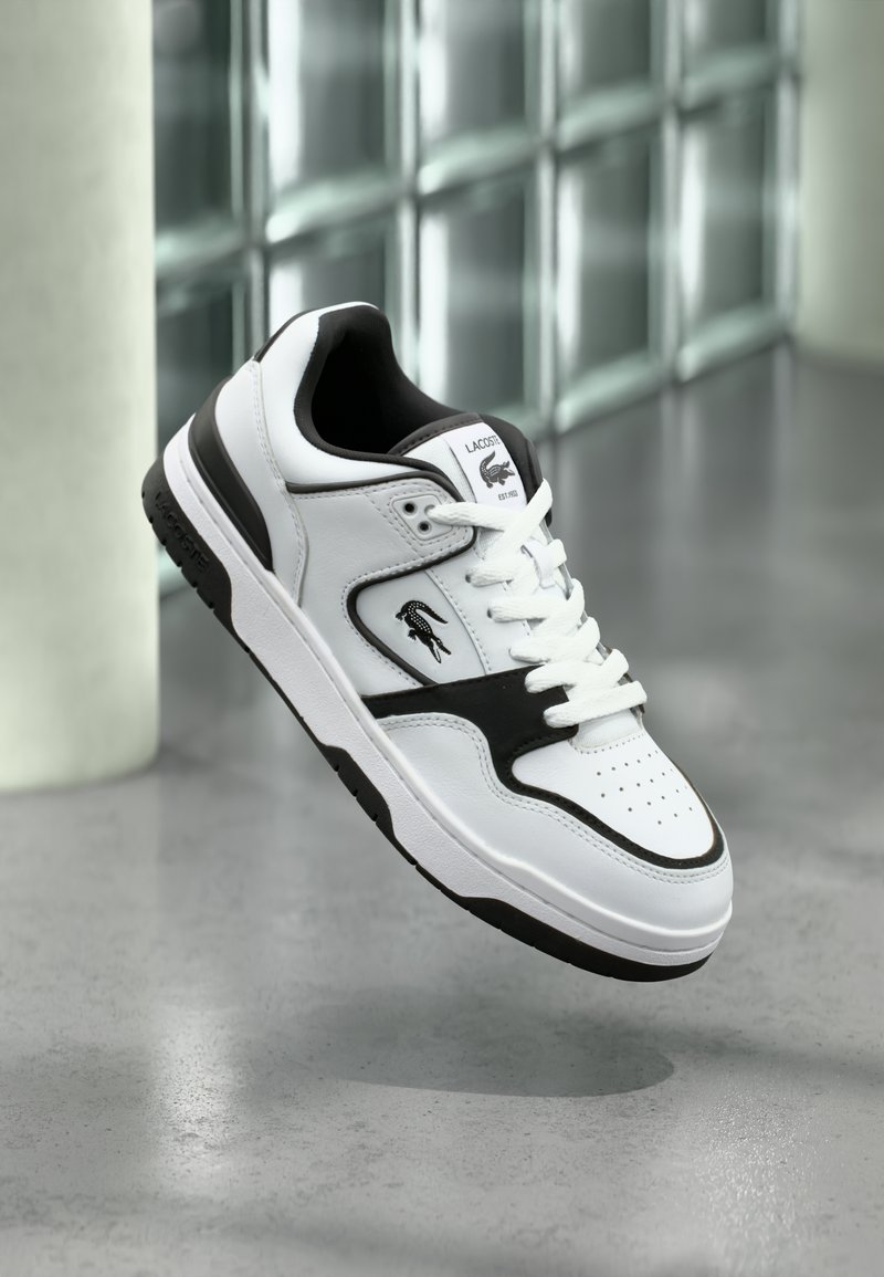 Λευκό και μαύρο αθλητικό παπούτσι Lacoste με κορδόνια, διάτρητο μπροστινό μέρος και λογότυπο στο πλάι και στη γλώσσα, εμφανίζεται να αιωρείται πάνω από γκρι πάτωμα.