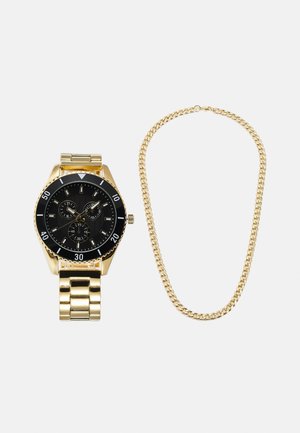 Relógio de ouro com mostrador preto e três submostradores; pulseira de metal dourada com elos. Colar de corrente dourada adjacente com um design de elos redondos.