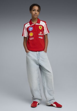 Jonge persoon draagt een rood Ferrari racehemd met sponsorlogo's, lichtblauwe losse jeans en rode Puma schoenen, staat met handen in de zakken.