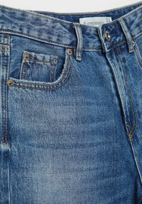 Detailný záber modrých riflí so zákazníckym vreckom vpredu, malým vreckom na drobné, putkami na opasok, kovovým gombíkom a viditeľnou veľkostnou štítkou "34".