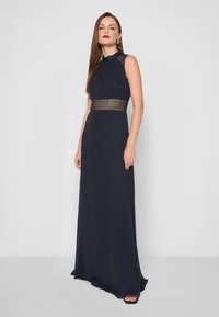 Námornícky modré maxi šaty so zberaným výstrihom, priesvitnými čipkovými detailmi a zložitými ozdobami v oblasti pása. Hladká, splývavá látka.
