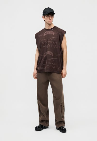 Νεαρός άνδρας που φοράει καφέ αμάνικο μπλουζάκι με γραφικά, καφέ παντελόνια cargo, μαύρο καπέλο και μαύρα παπούτσια, όρθιος και κοιτάζει στο πλάι.