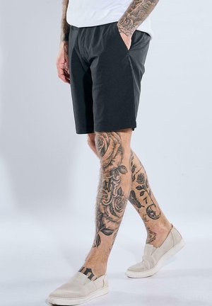 Mann trägt schwarze Shorts und beige Slip-on-Schuhe, zeigt detaillierte florale und Schrift-Tattoos, die beide Beine und Unterarme bedecken.