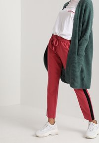Cardigan verde in maglia, t-shirt bianca, pantaloni jogger rossi con strisce laterali nere e sneakers bianche. Outfit casual con vestibilità comoda e contrasti di texture.